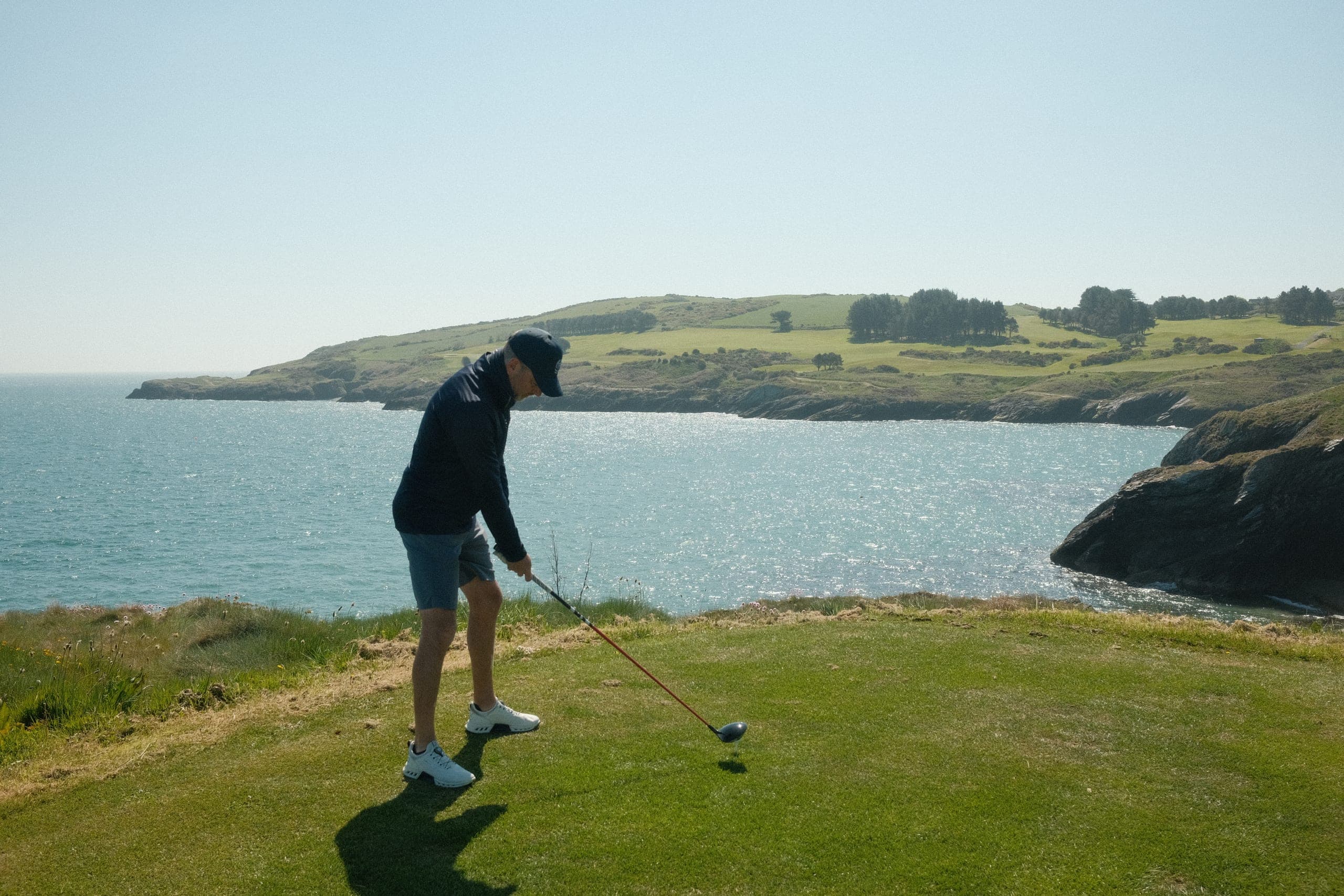 A golfer teeing off from a clifftop over a bright cove.