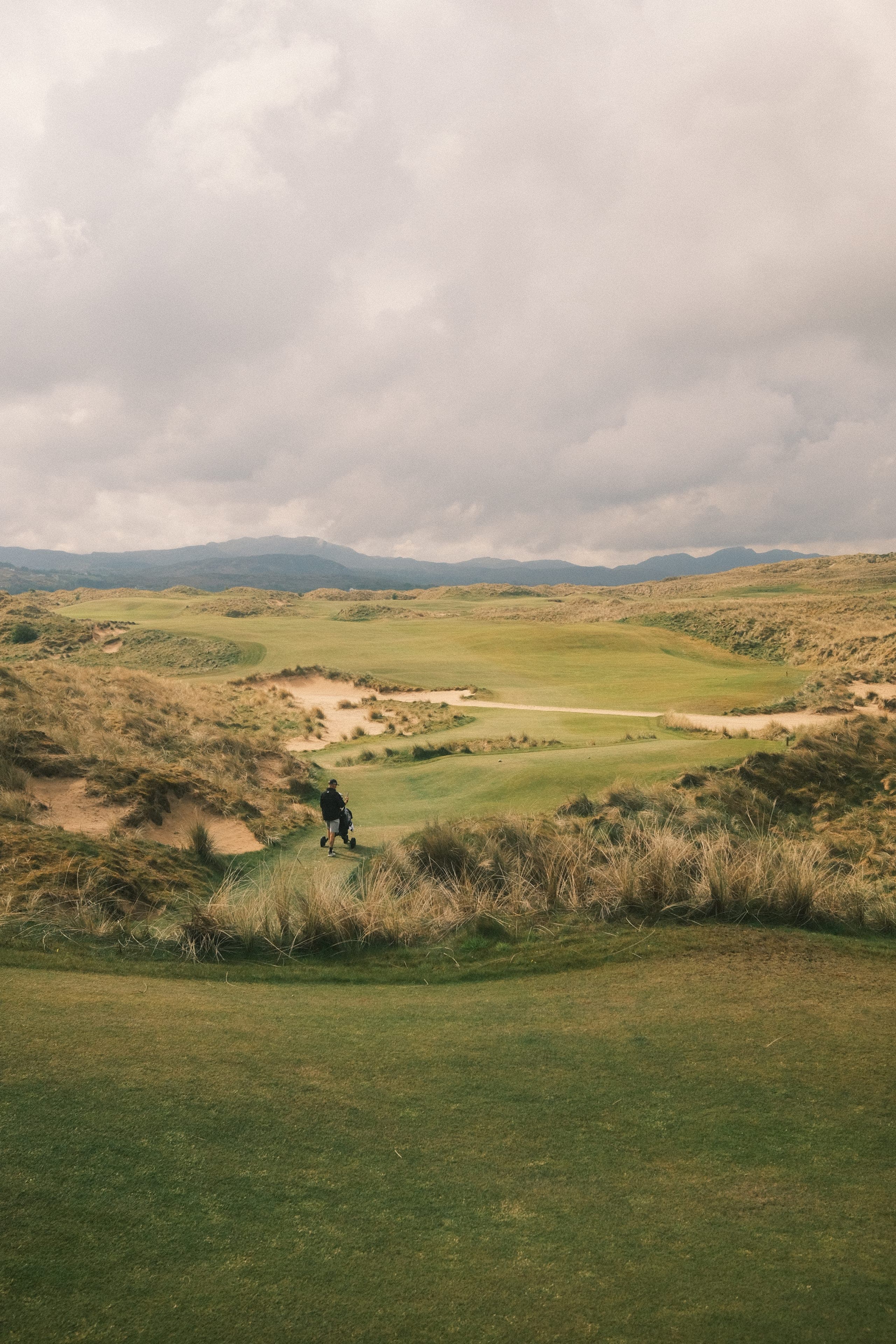 A golfer wheeling a push cart through a valley of high dunes.