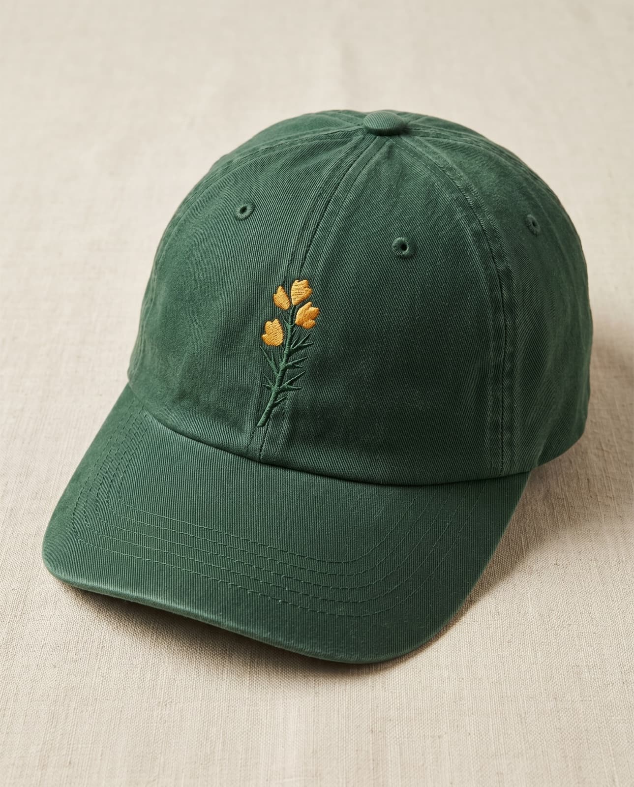 A washed racing-green cotton dad cap with a small green and gold embroidered gorse on the front and a raw brass buckle.