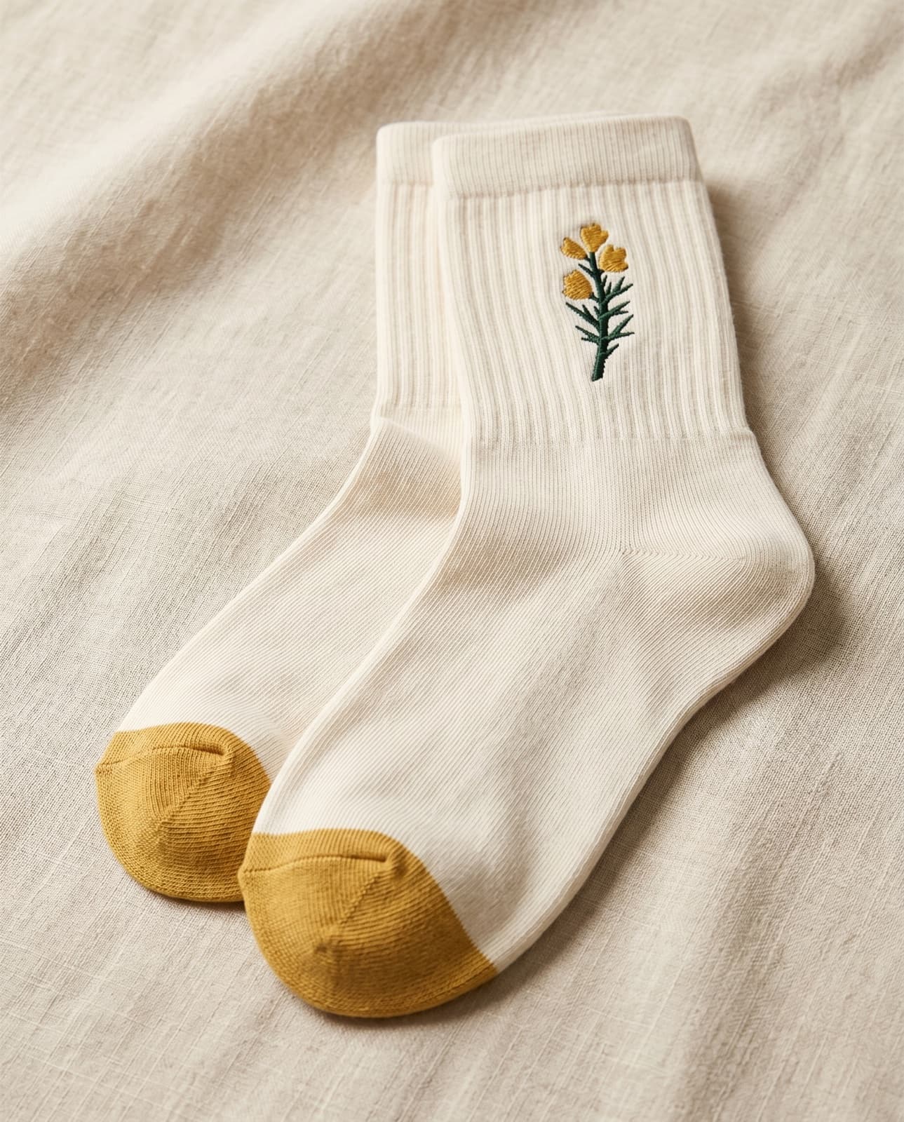 Cream cotton crew socks with a small embroidered gorse at the cuff and a solid gold toe, folded on cream linen.