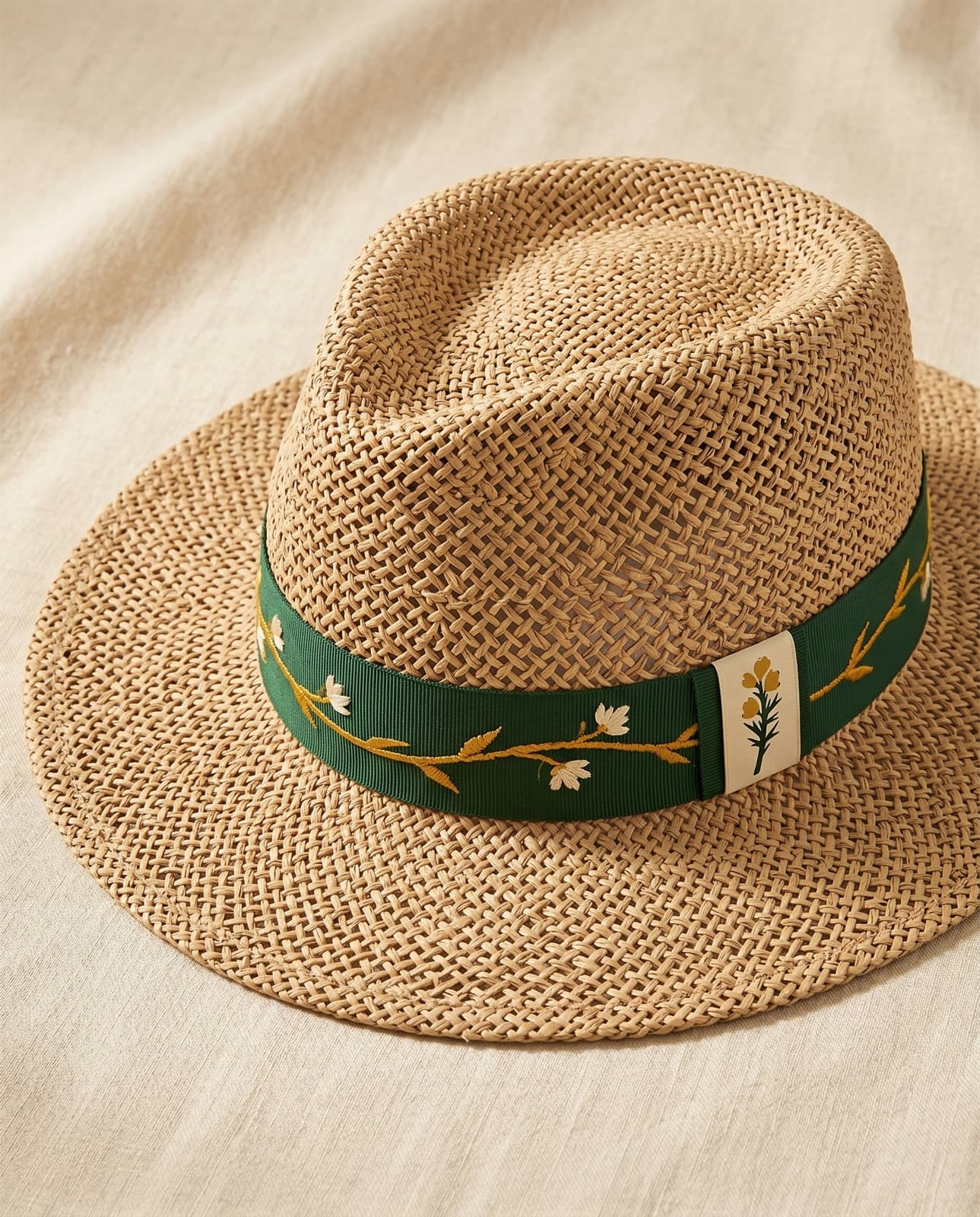 A natural straw hat with a flat brim and a racing-green band carrying a gold gorse vine, on cream linen.